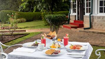 A table set outdoors with various breakfast items, including waffles, fruit, and beverages, in a garden setting with a building in the background.