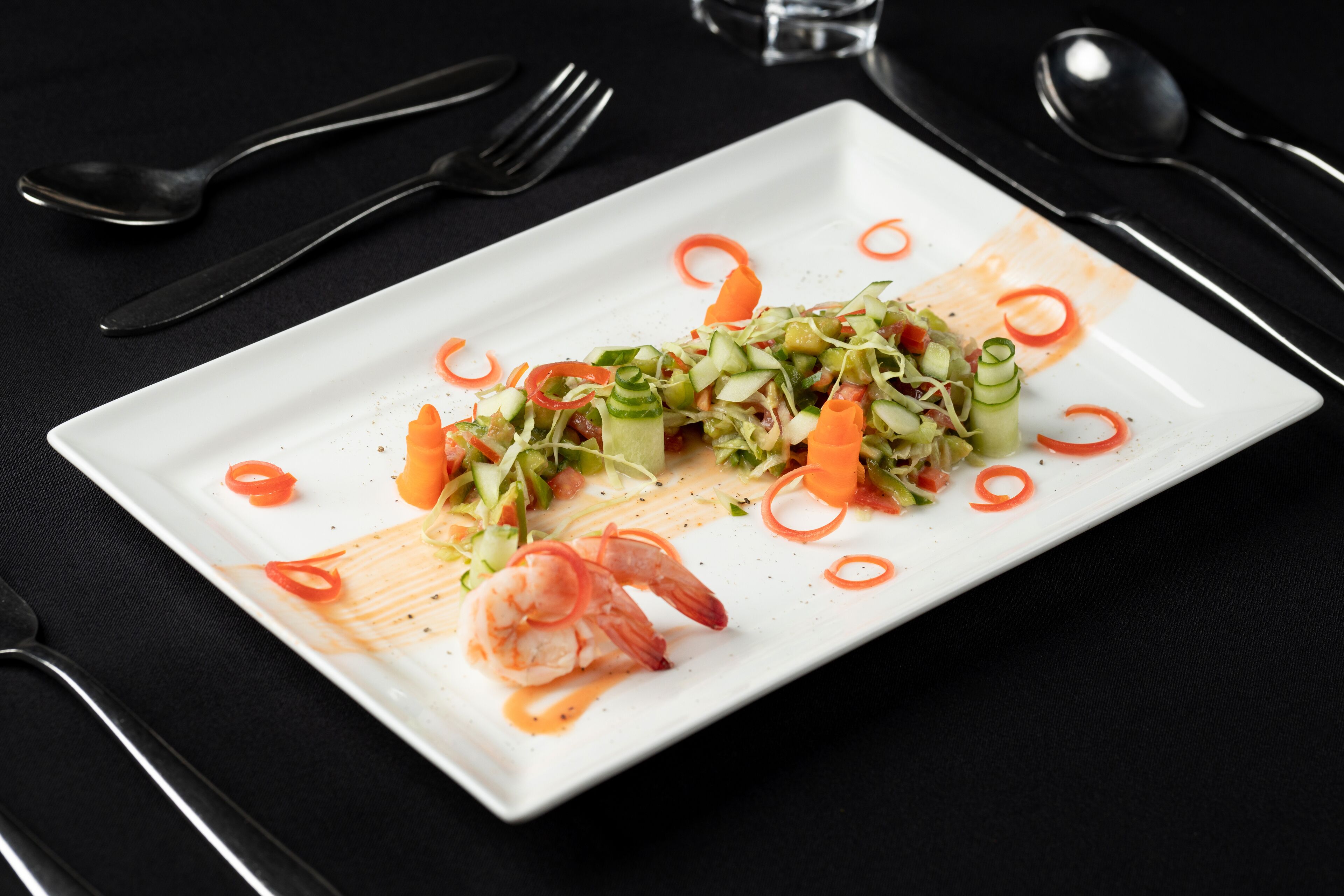 A white rectangular plate with a seafood dish, including shrimp and vegetables, on a black tablecloth.