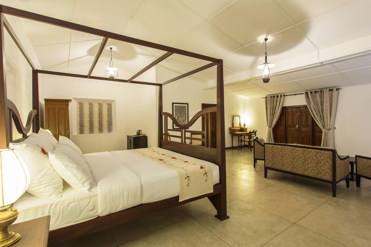 Una cama con dosel, una mesita de noche, una lámpara, una silla y un escritorio con otra silla en una habitación con suelo de baldosas y mobiliario de madera.