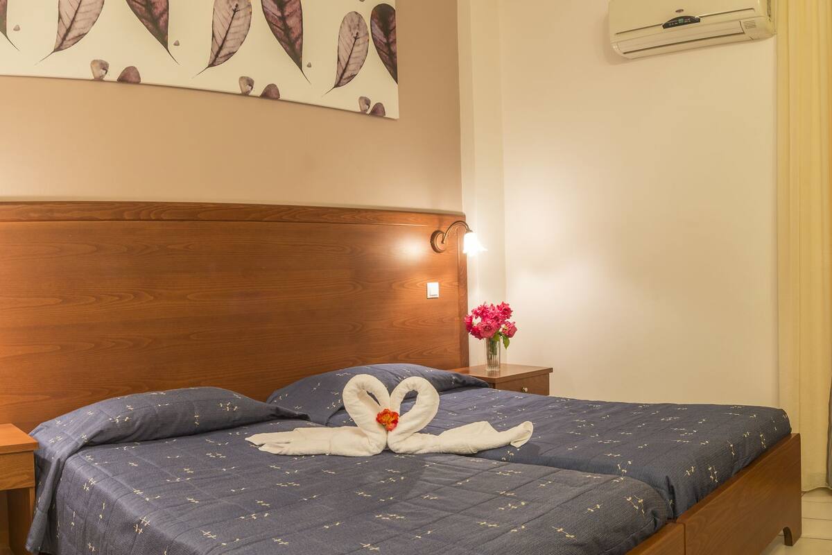 A hotel room with a wooden headboard, a bed with a blue bedspread and a heart-shaped towel decoration, a wooden nightstand with a phone, a vase with flowers, a fruit bowl on a wooden table, and a wall-mounted air conditioner.