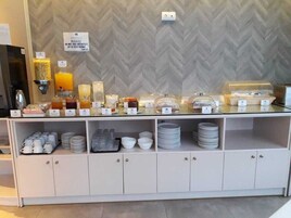 Um buffet de café da manhã com diversos alimentos e bebidas, uma placa indicando para não levar o café da manhã para o quarto, e pratos e xícaras organizados.