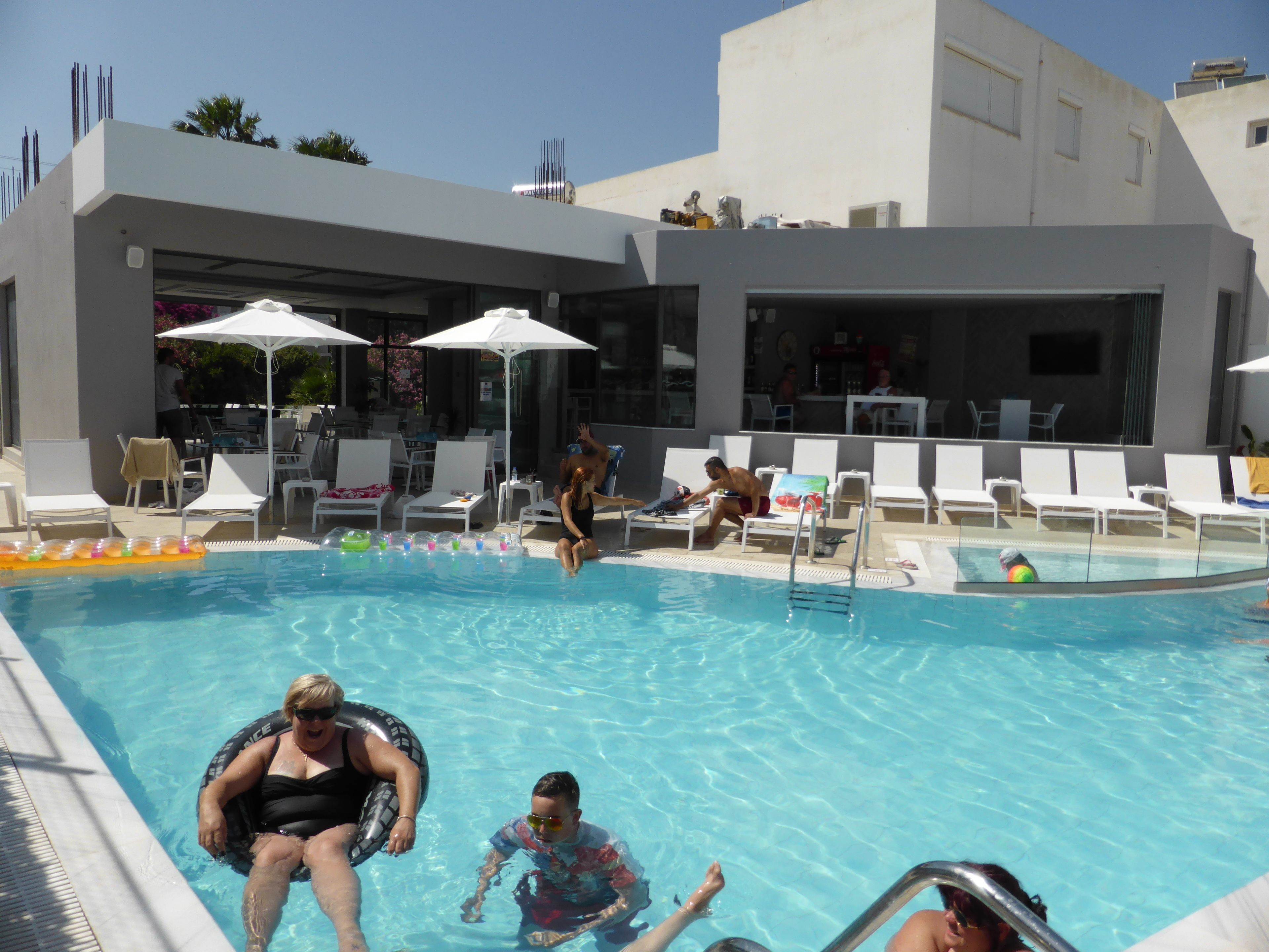 A swimming pool with lounge chairs and umbrellas, a bar area, and a modern building in the background.
