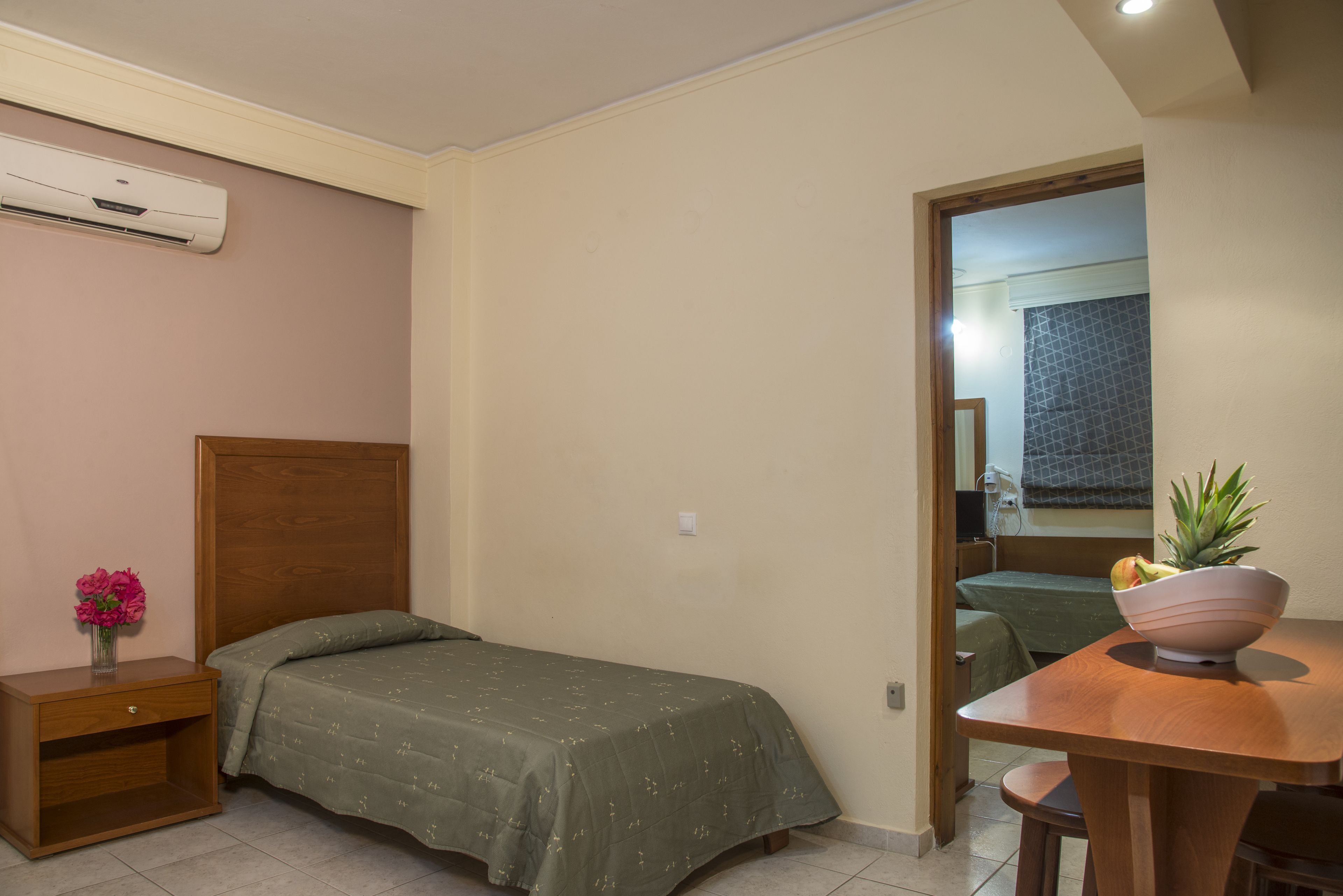 Un dormitorio pequeño y ordenado con una cama individual, una mesita de noche con un florero y una mesa de madera con un frutero.