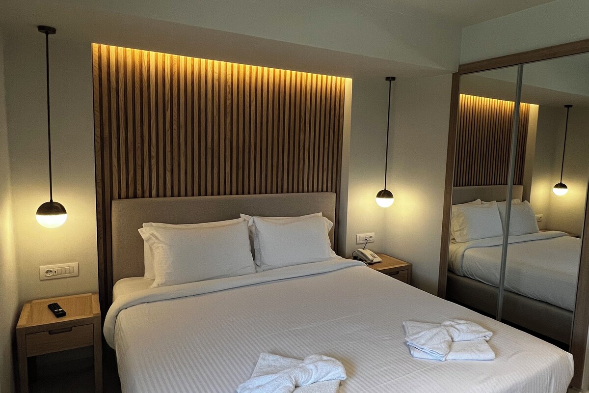 A modern hotel room with a large bed, a wooden headboard, bedside tables with lamps, and a full-length mirror.