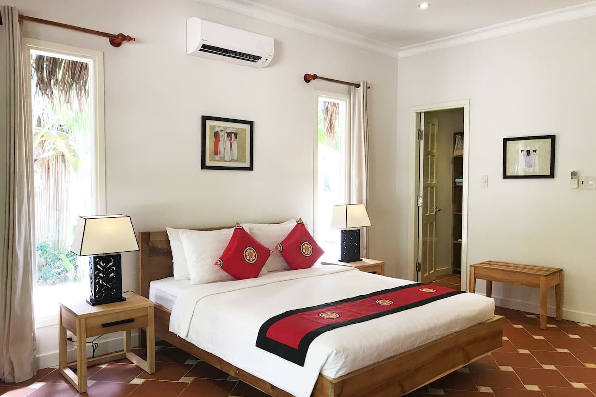 A neatly arranged bedroom with a wooden bed, two bedside tables, a nightstand, a red throw blanket, and a wall-mounted air conditioner.