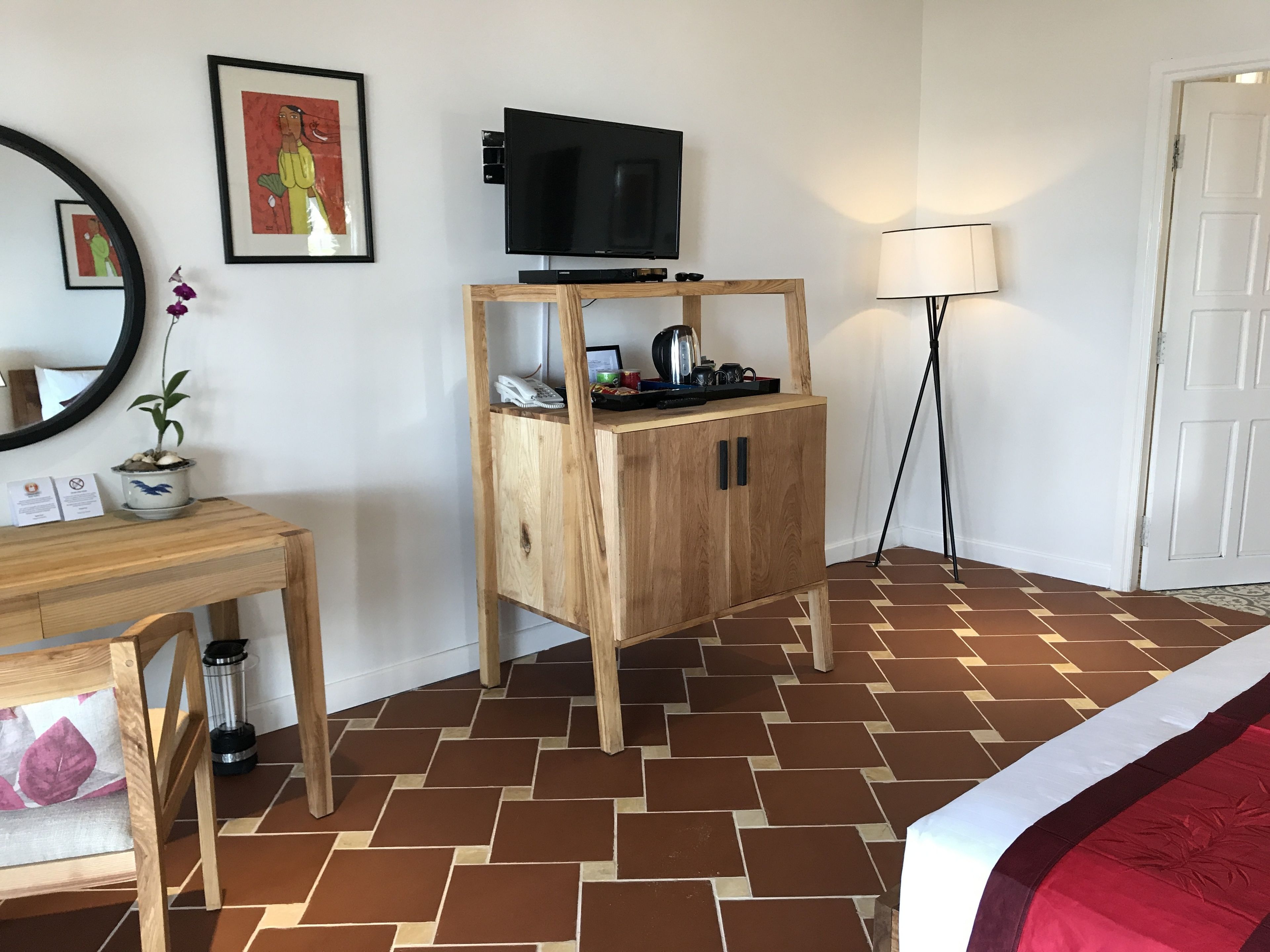 Habitación de hotel con escritorio de madera, televisor en un mueble bajo, mesita de noche, lámpara y una puerta.