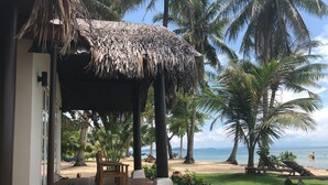 A beachside patio with a thatched roof, palm trees, a wooden bench, and a view of the sea. - Peppercorn Beach (Phu Quoc)