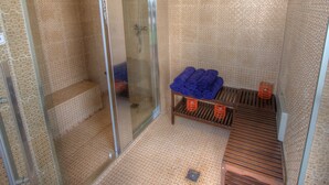 Una habitación con una puerta de vidrio que da a un área de ducha, una cama con una manta azul y dos cestas naranjas sobre un estante de madera.