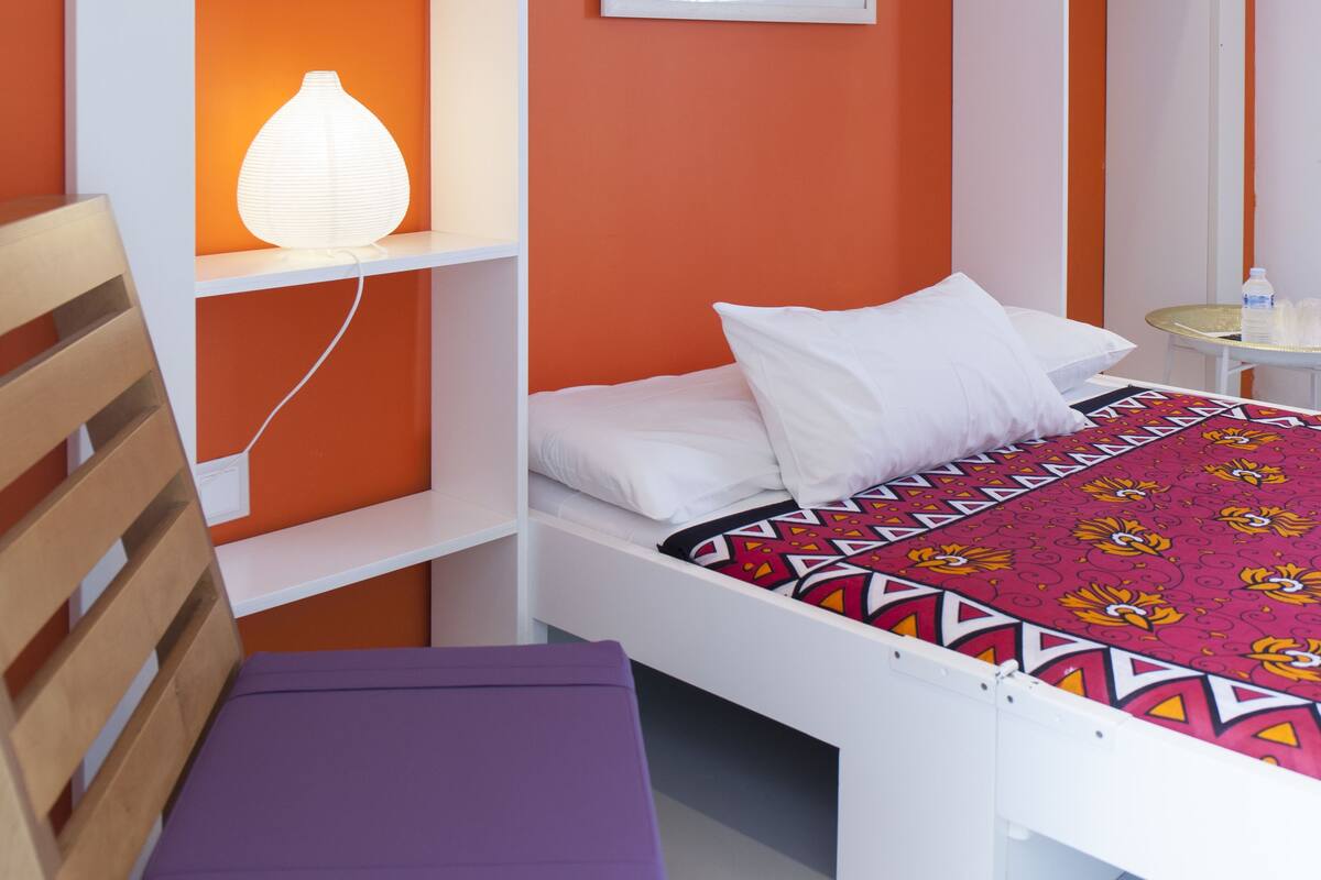 Una habitación compacta con una cama, un banco y una repisa con lámpara.