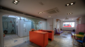 Un interior moderno con un mostrador de recepción naranja brillante, separadores de vidrio y vista a un coche rojo en el exterior.