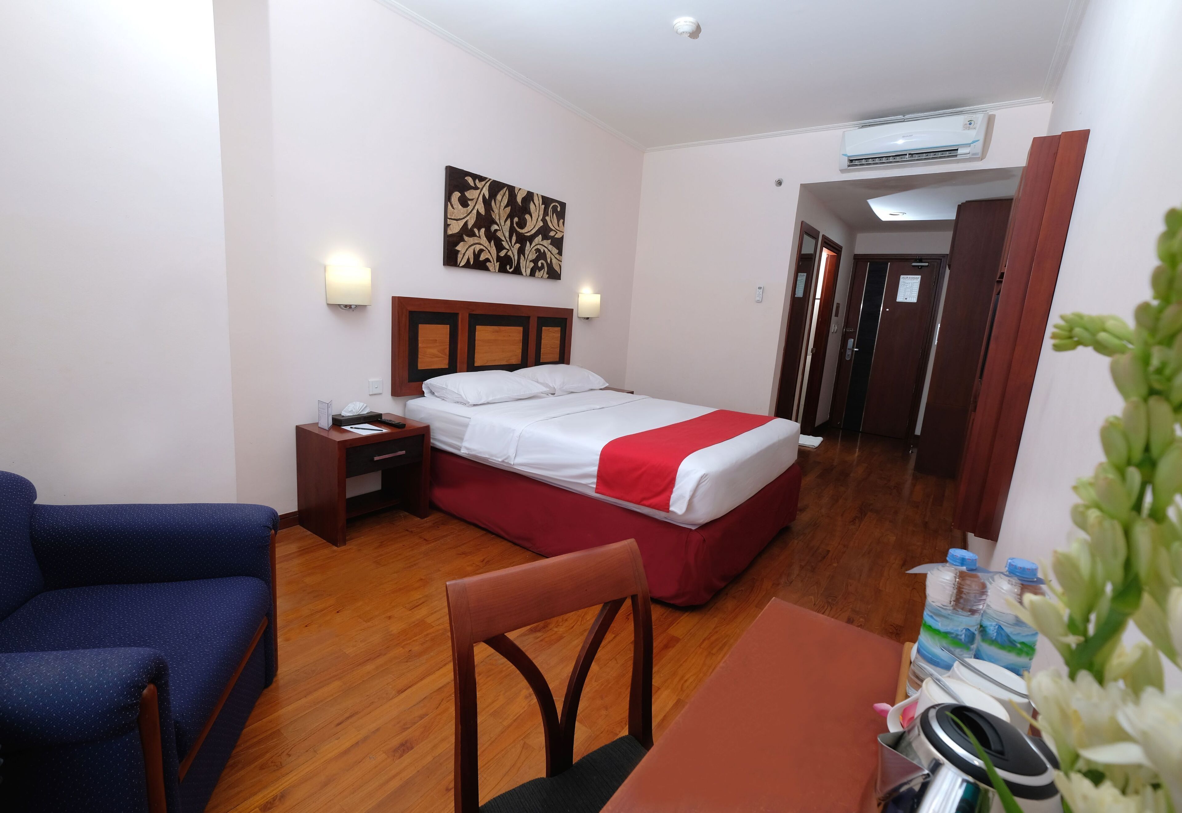 A hotel room with a bed, a desk, a chair, a nightstand, and a wall-mounted air conditioner.
