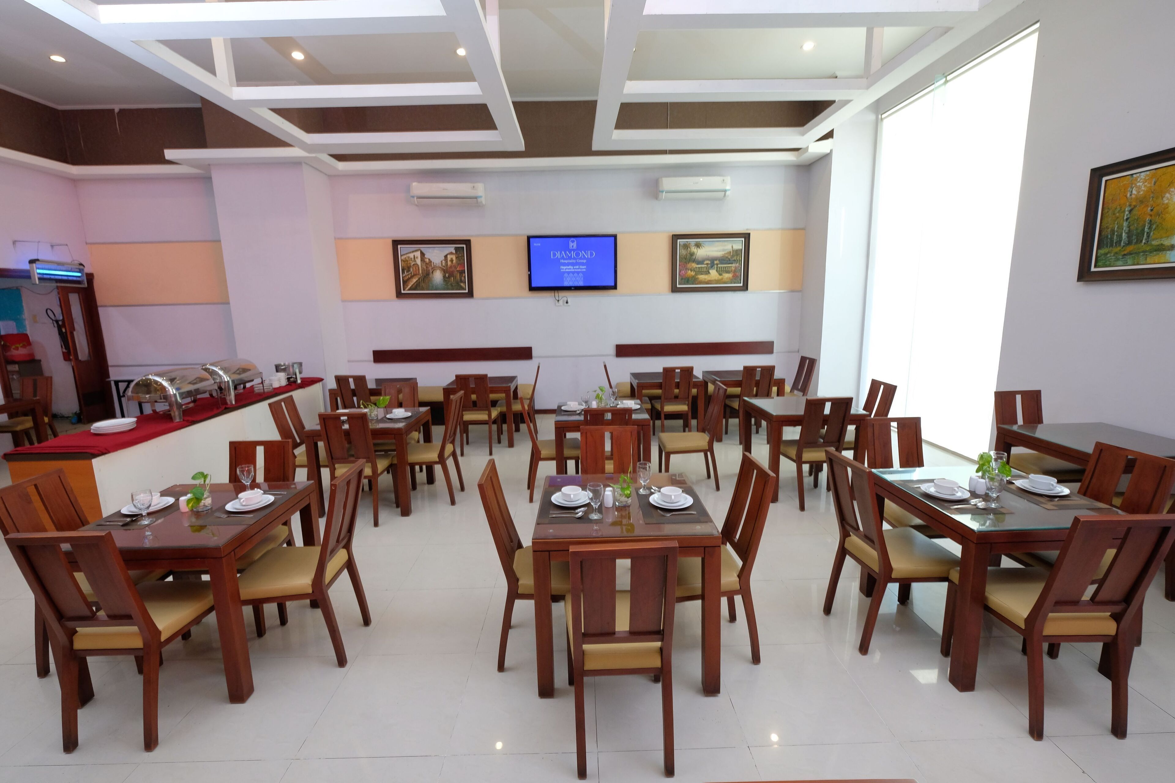 Un comedor de hotel con varias mesas y sillas de madera, una pantalla de televisión en la pared y un mostrador de buffet al fondo.