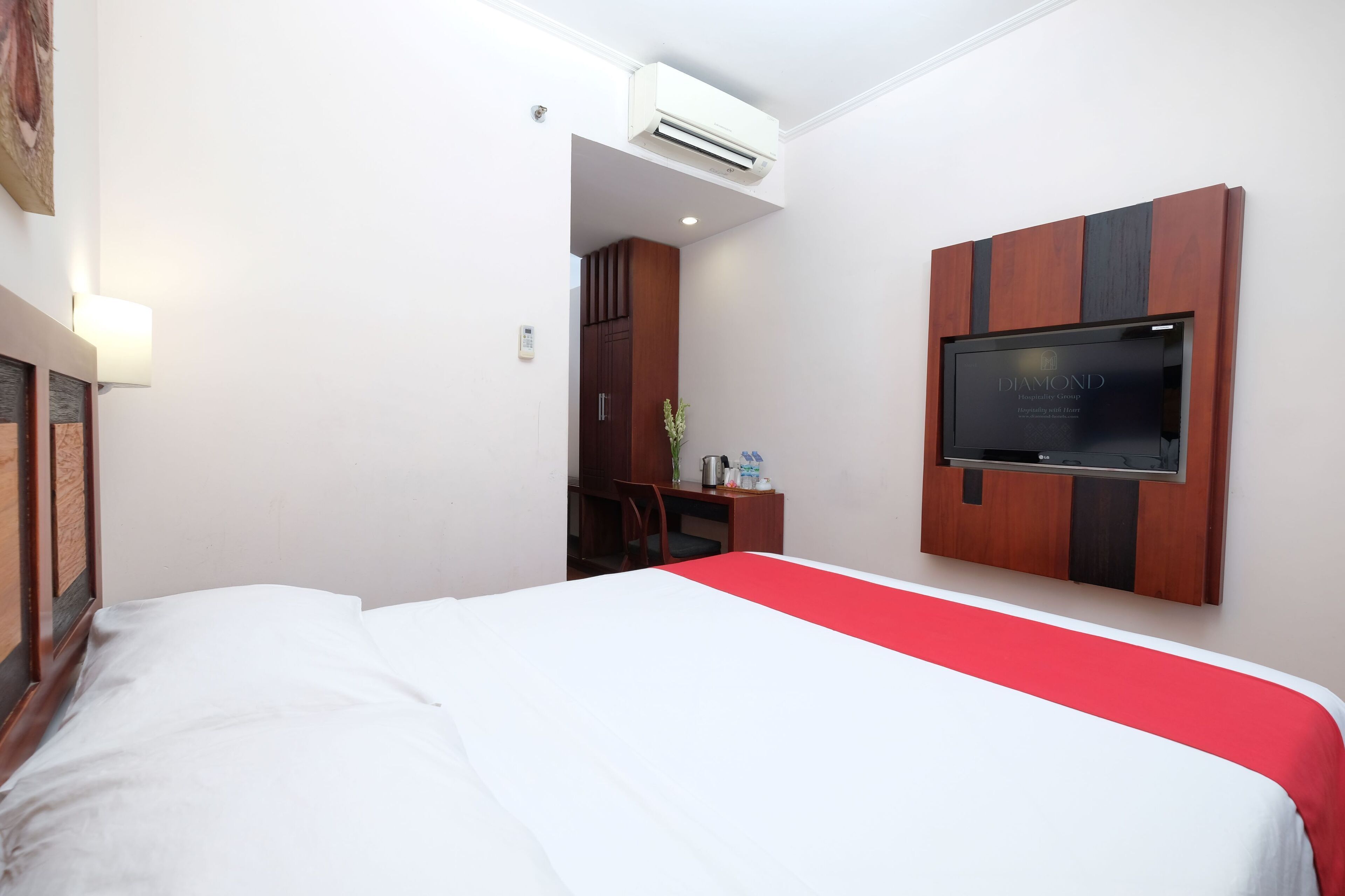 A hotel room with a bed, a desk, a chair, a TV mounted on the wall, and an air conditioning unit.