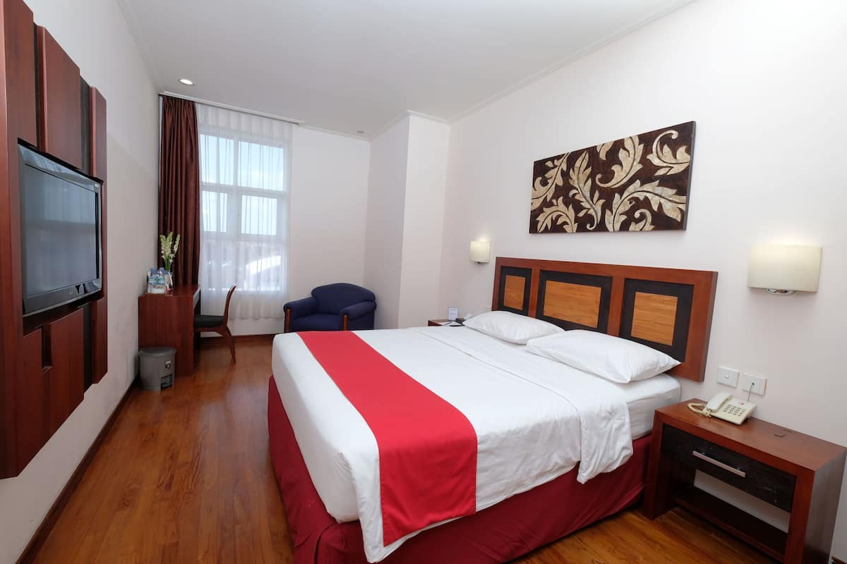 A hotel room with a large bed, a wooden nightstand, a desk, a chair, a TV, and a window with curtains.
