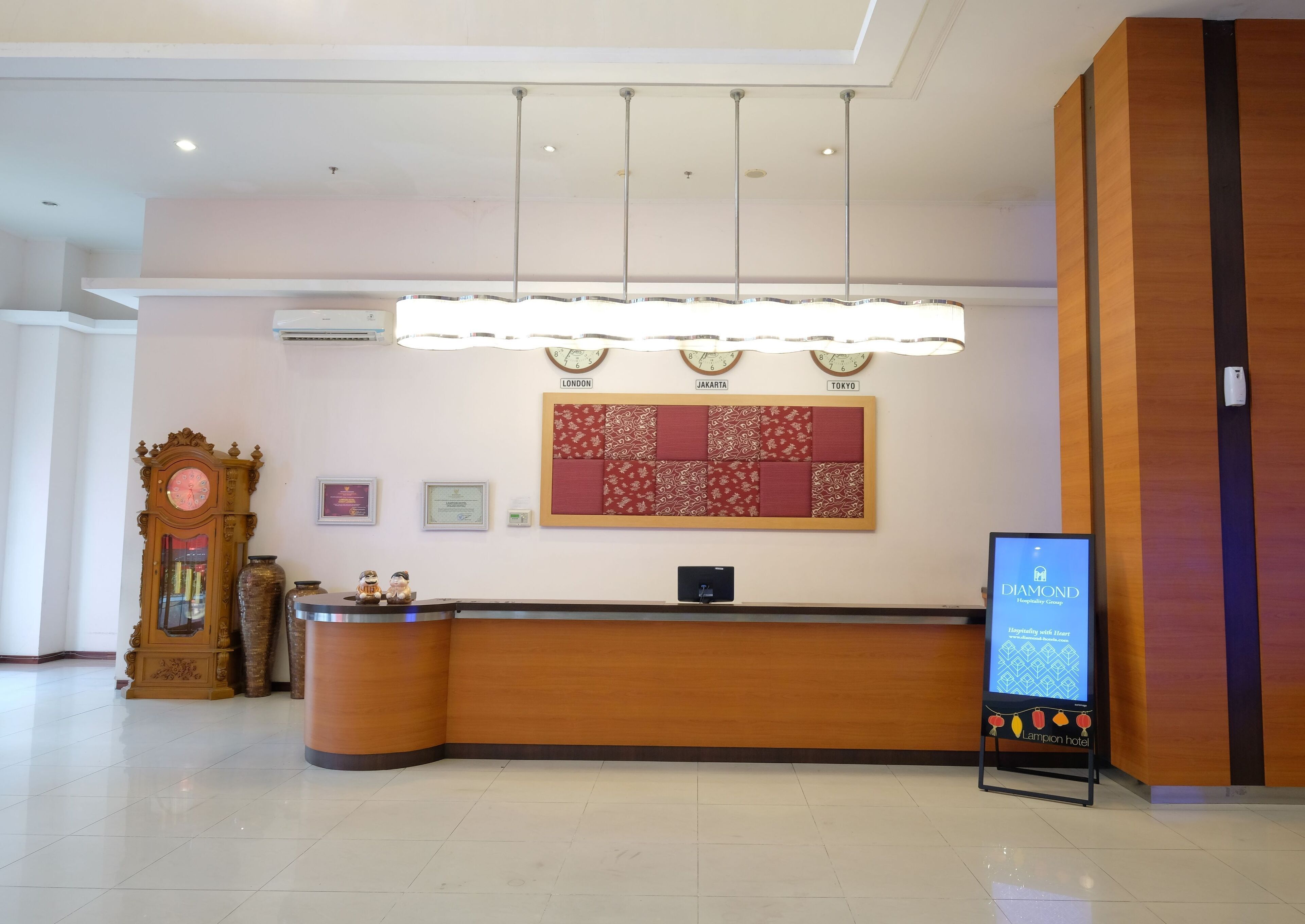 Zona de recepción de hotel con un mostrador de madera, un reloj de pared, un certificado enmarcado y un panel de anuncios digital.