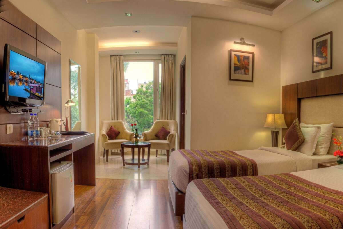 A hotel room with two beds, a TV, a desk with a refrigerator, a chair, and a view of the outdoors.