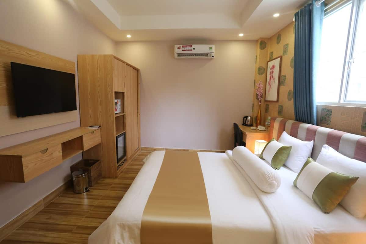 A hotel room with a bed, a TV mounted on the wall, a wardrobe, an air conditioning unit, and a window with curtains.