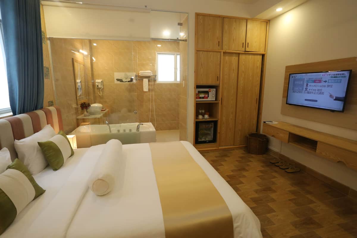 A hotel room with a large bed, a TV, wooden cabinets, a small TV, and a bathroom with a glass shower enclosure.
