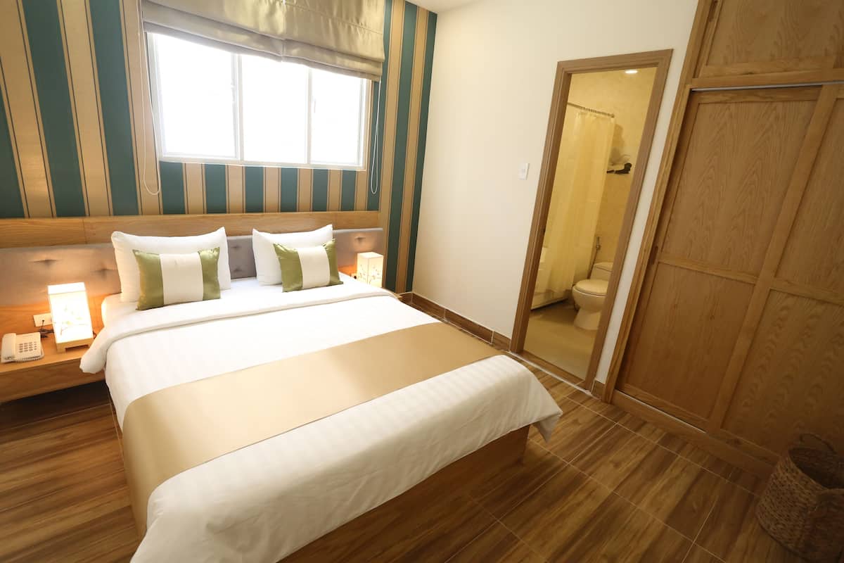 A hotel room with a large bed, bedside tables, a window with curtains, a wooden floor, and a wooden door.