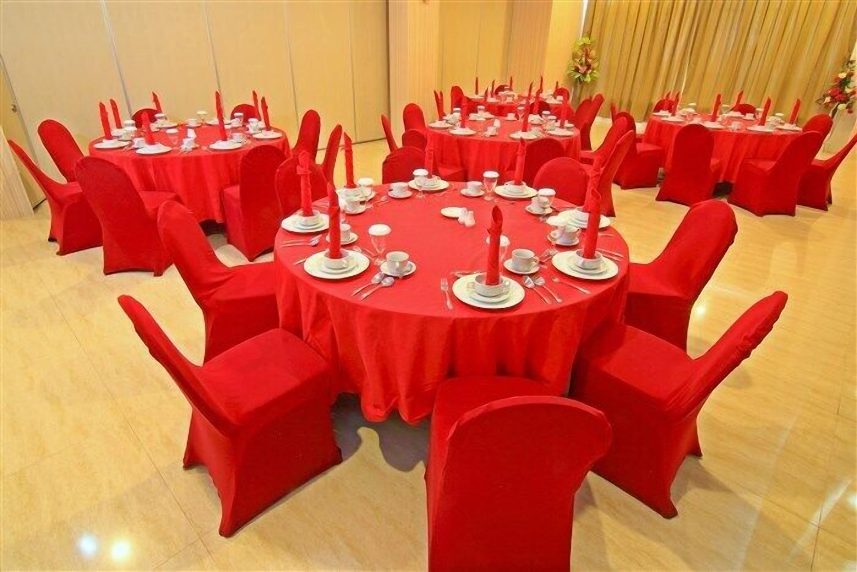 Un salón de banquetes con mesas redondas cubiertas con manteles rojos, cada una con platos blancos, cubiertos y servilletas rojas.