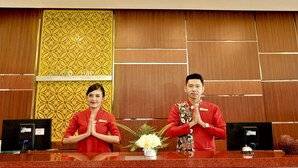 Two receptionists at a hotel reception desk with a decorative panel and a nameplate in the background. - Grand HAP Hotel (Surakarta)