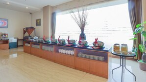 A buffet setup with various food containers and serving utensils, a large window with curtains, and a potted plant. - Grand HAP Hotel (Surakarta)