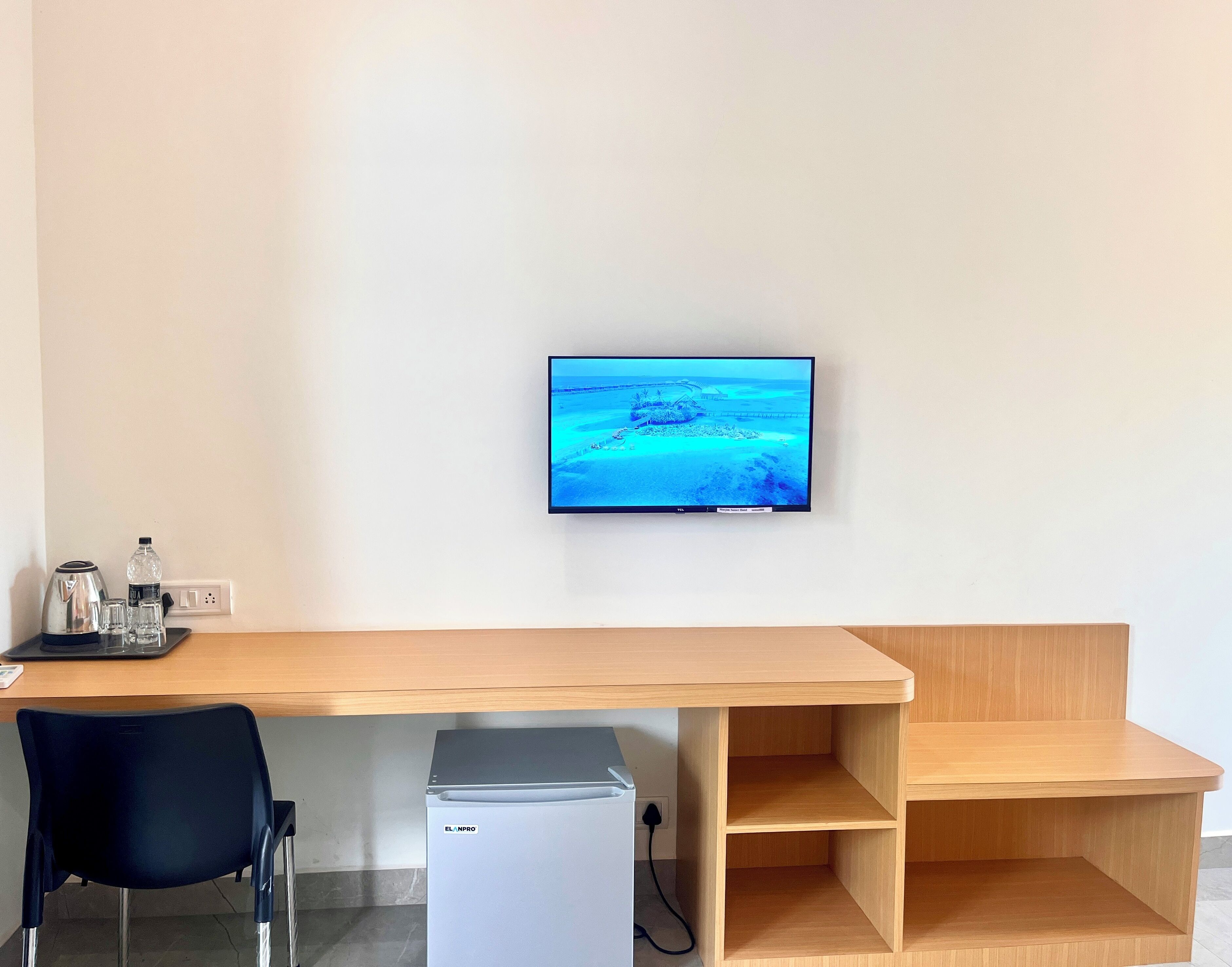 A modern room with a wooden desk, a black chair, a small refrigerator, and a flat-screen TV mounted on the wall displaying a beach scene.