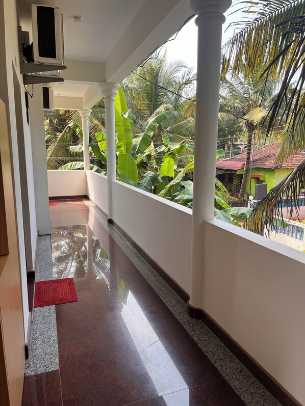 A corridor with a polished floor, white pillars, and a view of a green house and palm trees. - Morjim Sunset Hotel - 300 Meter to Morjim Beach with Pool & Kitchen Apartments (Morjim)