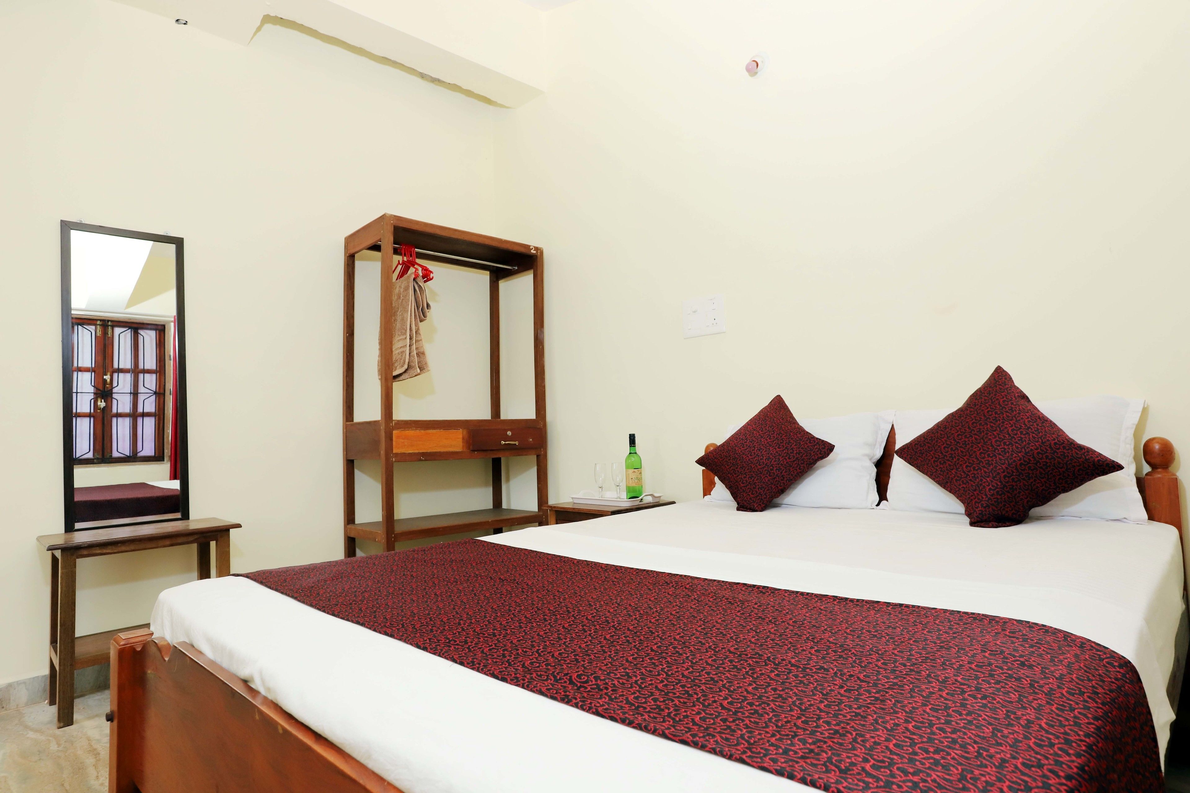 A hotel room with a large bed, a wooden wardrobe, a bedside table with a wine bottle, and a mirror on the wall.