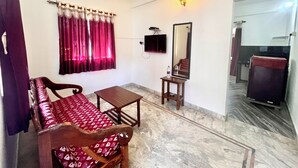 A room with a sofa, a small table, a ceiling fan, a TV, a mirror, and a door leading to another room. - Morjim Sunset Hotel (Morjim)