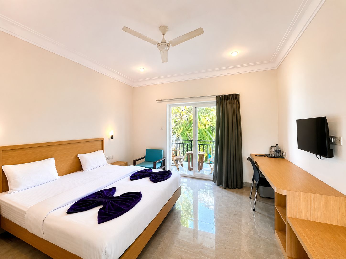 A hotel room with a bed, a desk, a chair, a TV, and a balcony with a view of palm trees.