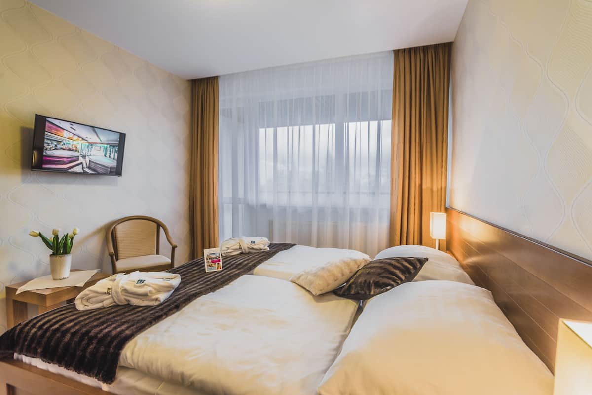 Una habitación de hotel con una cama grande, una silla, una mesita de noche con lámpara, un televisor en la pared y una ventana con cortinas.