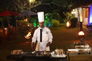 A chef grilling food outdoors at night with a fire pit and dining area in the background. - Orient Hotel (Bandarawela)