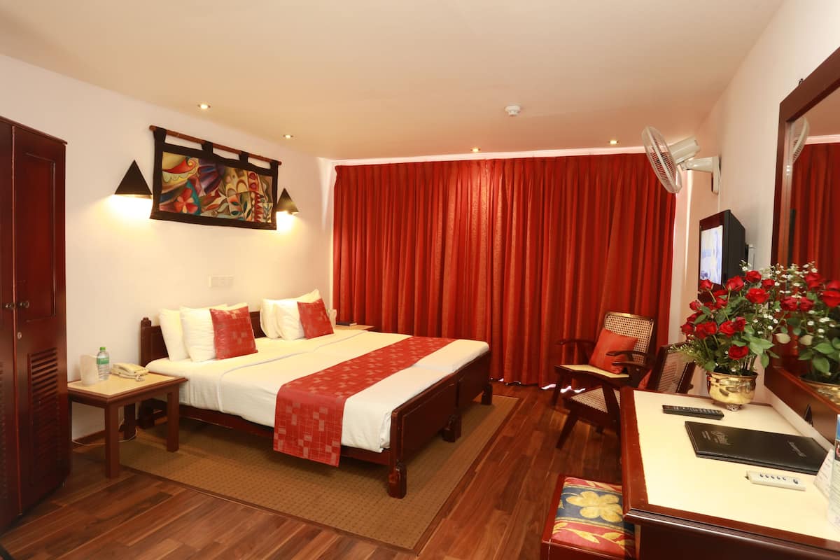 A hotel room with a large bed, a desk with a computer, a chair, a small table with a phone, a vase with flowers, and a painting on the wall.