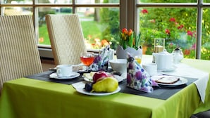 Une table de petit-déjeuner dressée avec une variété d’aliments, notamment des fruits, du pain et un verre de jus, et offrant une vue sur un jardin grâce à de grandes fenêtres.