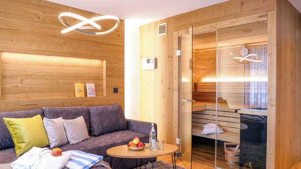 Une pièce moderne avec un mur en bois, un canapé gris, une petite table ronde ornée de fruits et de verres, et une porte en verre donnant sur un espace sauna.