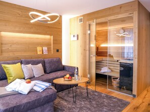 Quarto moderno com parede de madeira, sofá cinza, mesa redonda pequena com frutas e copos, e porta de vidro que dá para uma área de sauna.