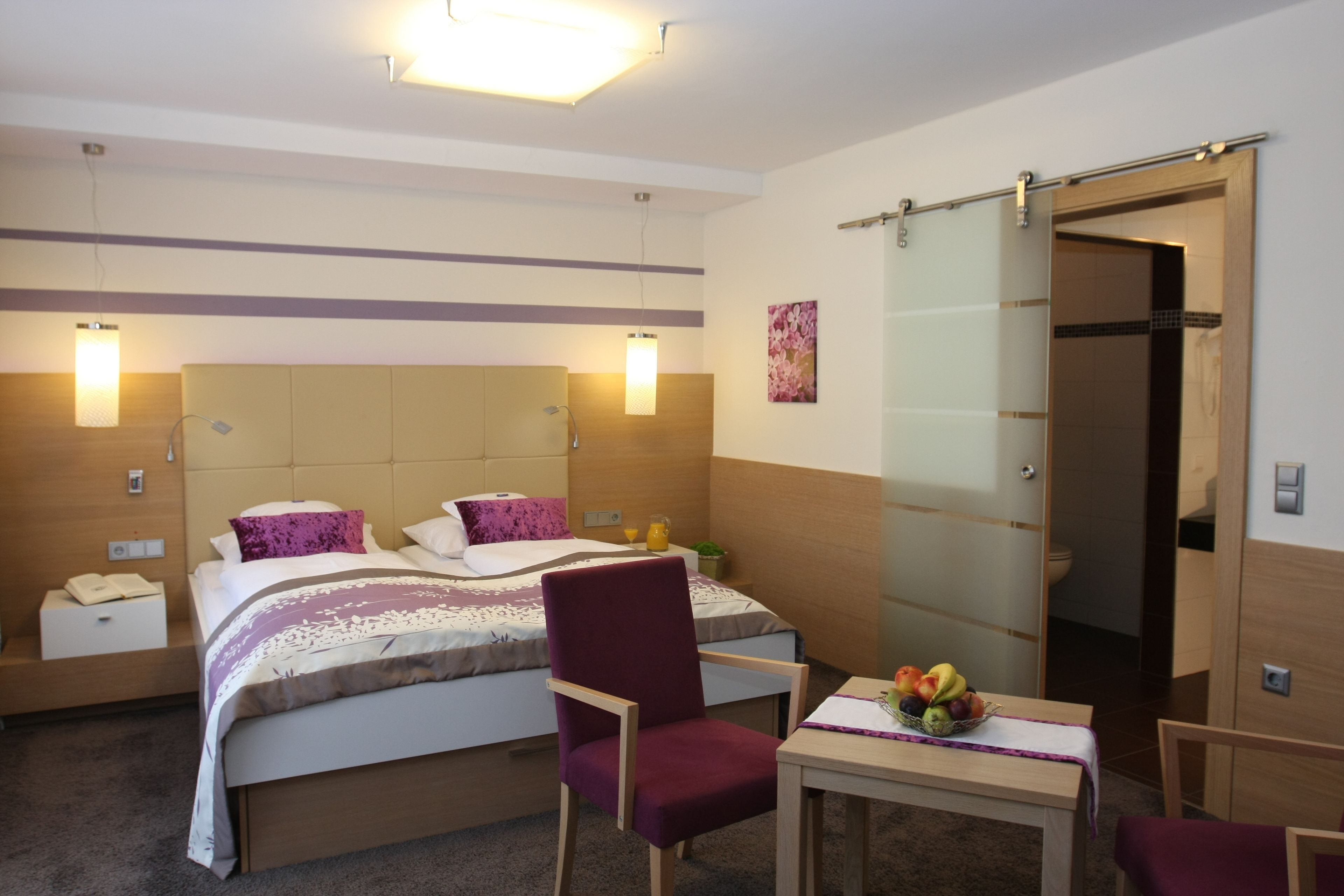 A hotel room with a bed, a chair, a table with fruit, and a bathroom door.
