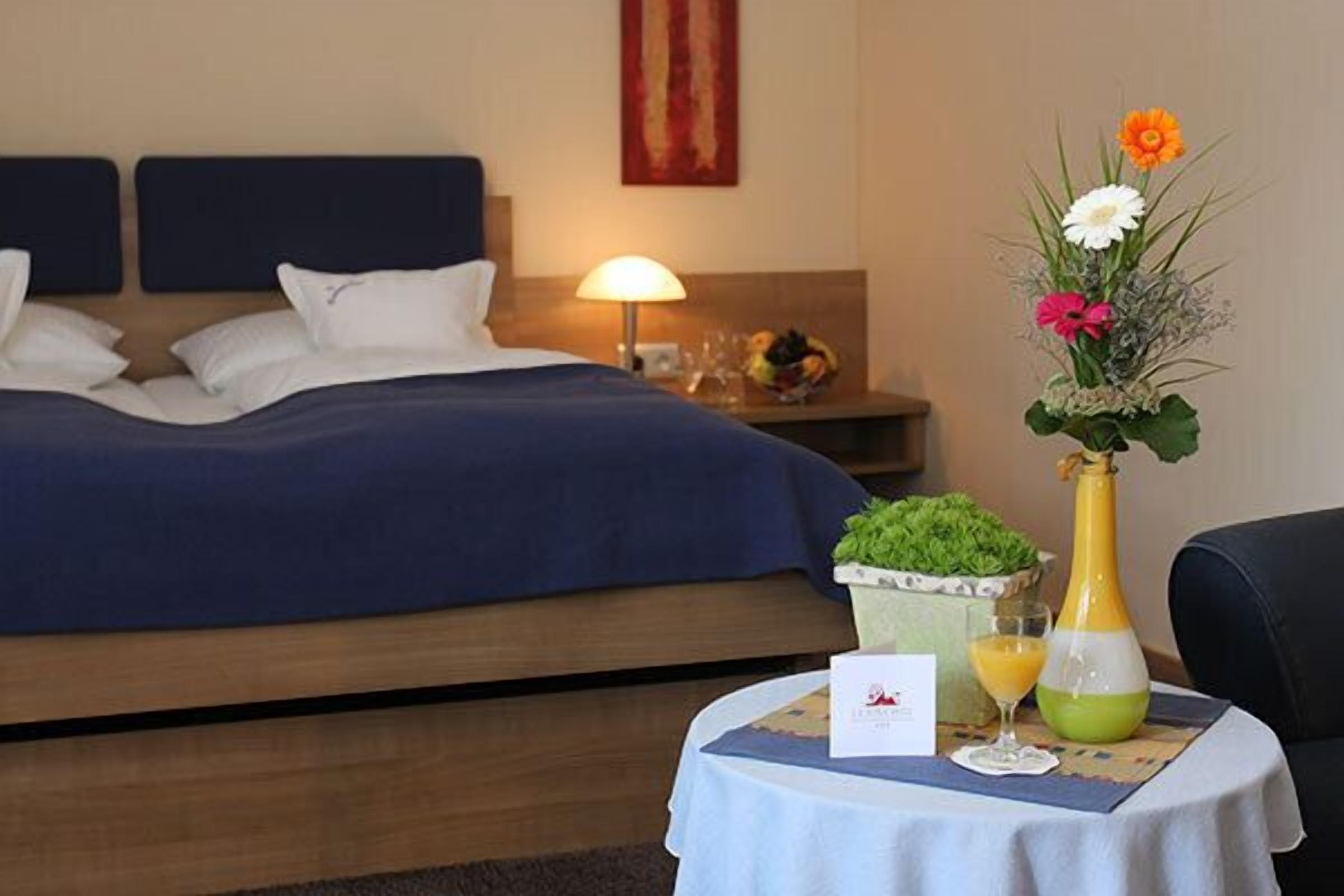 A hotel room with a bed, a nightstand with a lamp, a small table with a vase of flowers, a fruit bowl, and a bottle of juice.