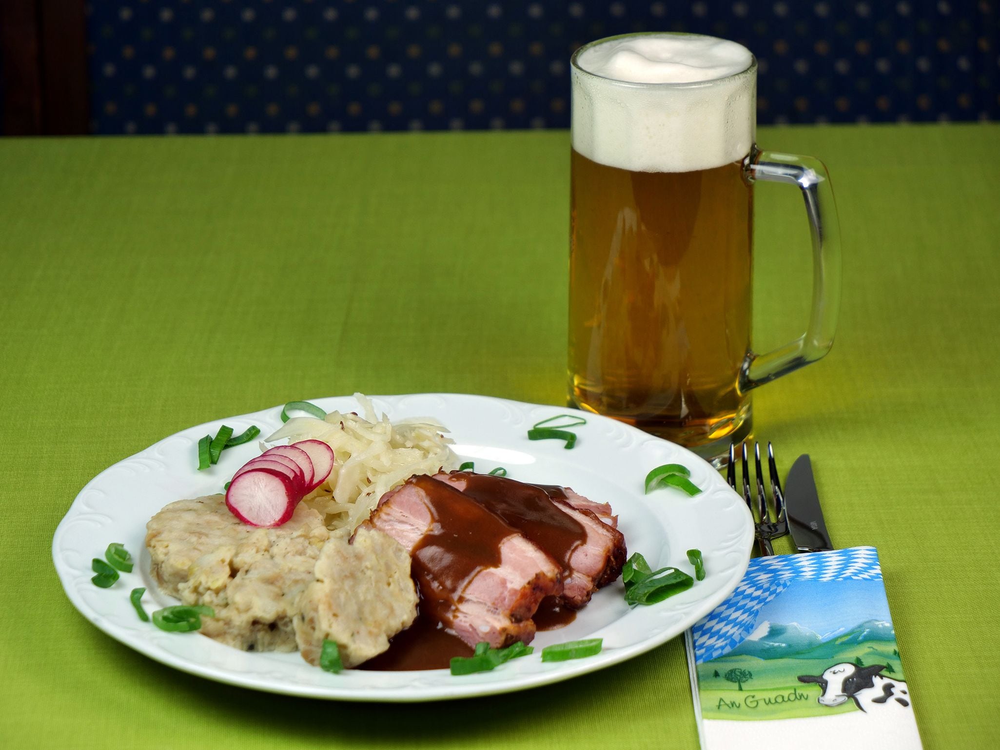 Une assiette avec des tranches de viande, de la choucroute et des radis, un verre de bière et une serviette en papier ornée d’une illustration de vache.