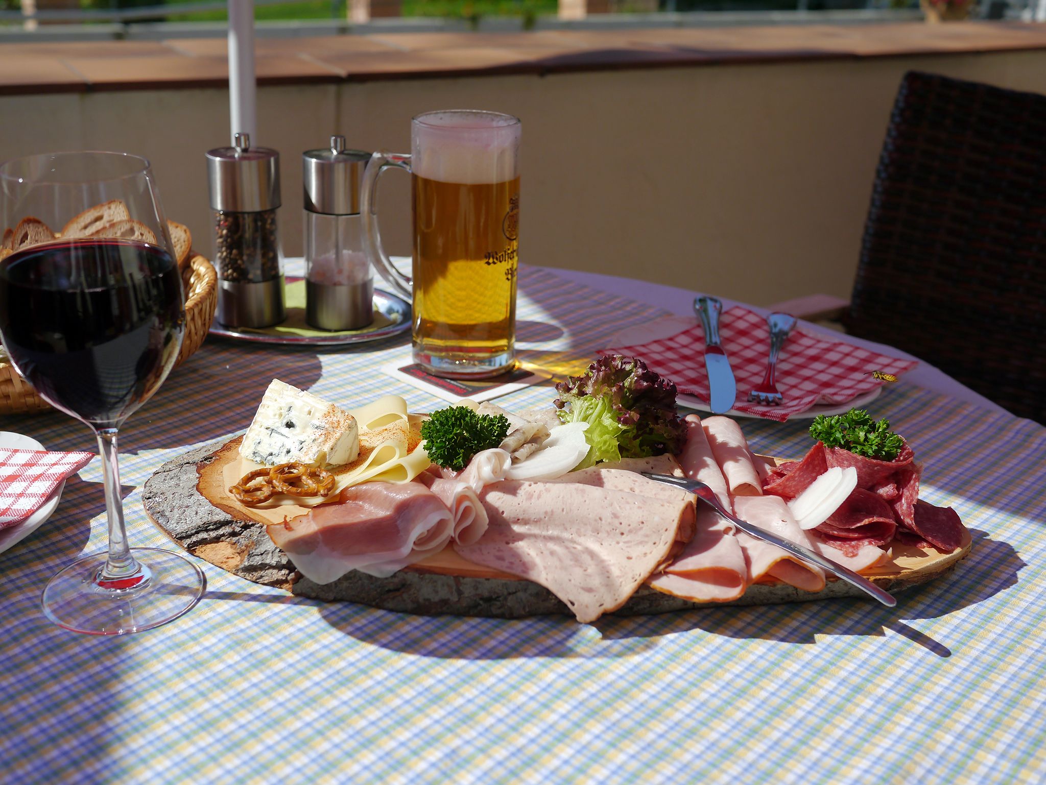 Una mesa con mantel a cuadros, una copa de vino tinto, una jarra de cerveza y una variedad de carnes y quesos.