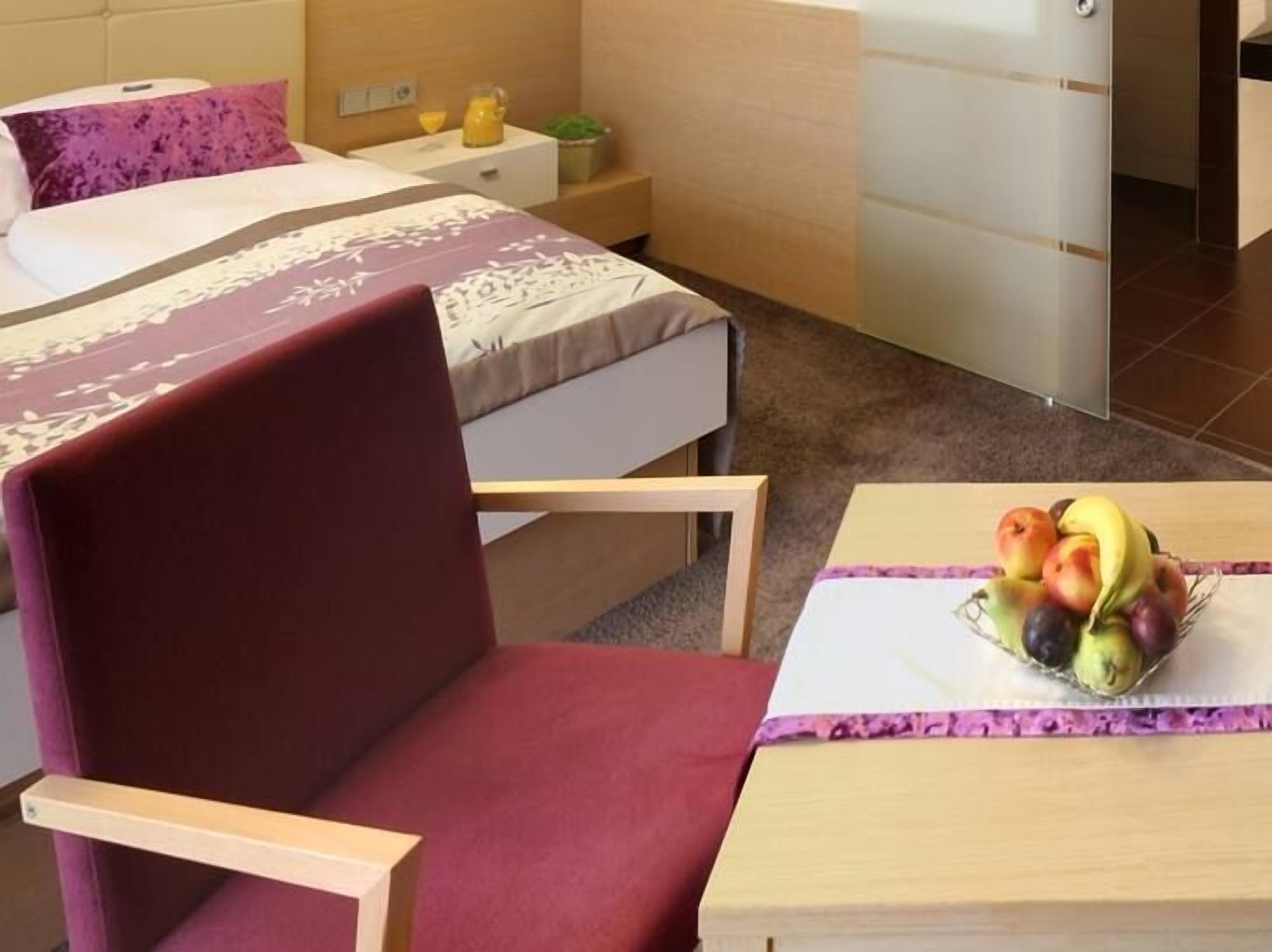 A hotel room with a bed, a chair, a small table with fruit, and a nightstand with a lamp and a plant.