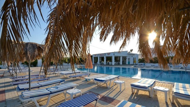 A pool area with lounge chairs and umbrellas, a clear sky, and a building in the background. - Nikos 2 Studios & Apartments (Kos)