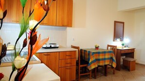 A kitchen with wooden cabinets, a dining table with a checkered tablecloth, and a small desk with a lamp. - Nikos 2 Studios & Apartments (Kos)