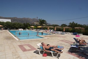 A pool area with multiple sun loungers and umbrellas, a swimming pool, and a mountainous backdrop.
