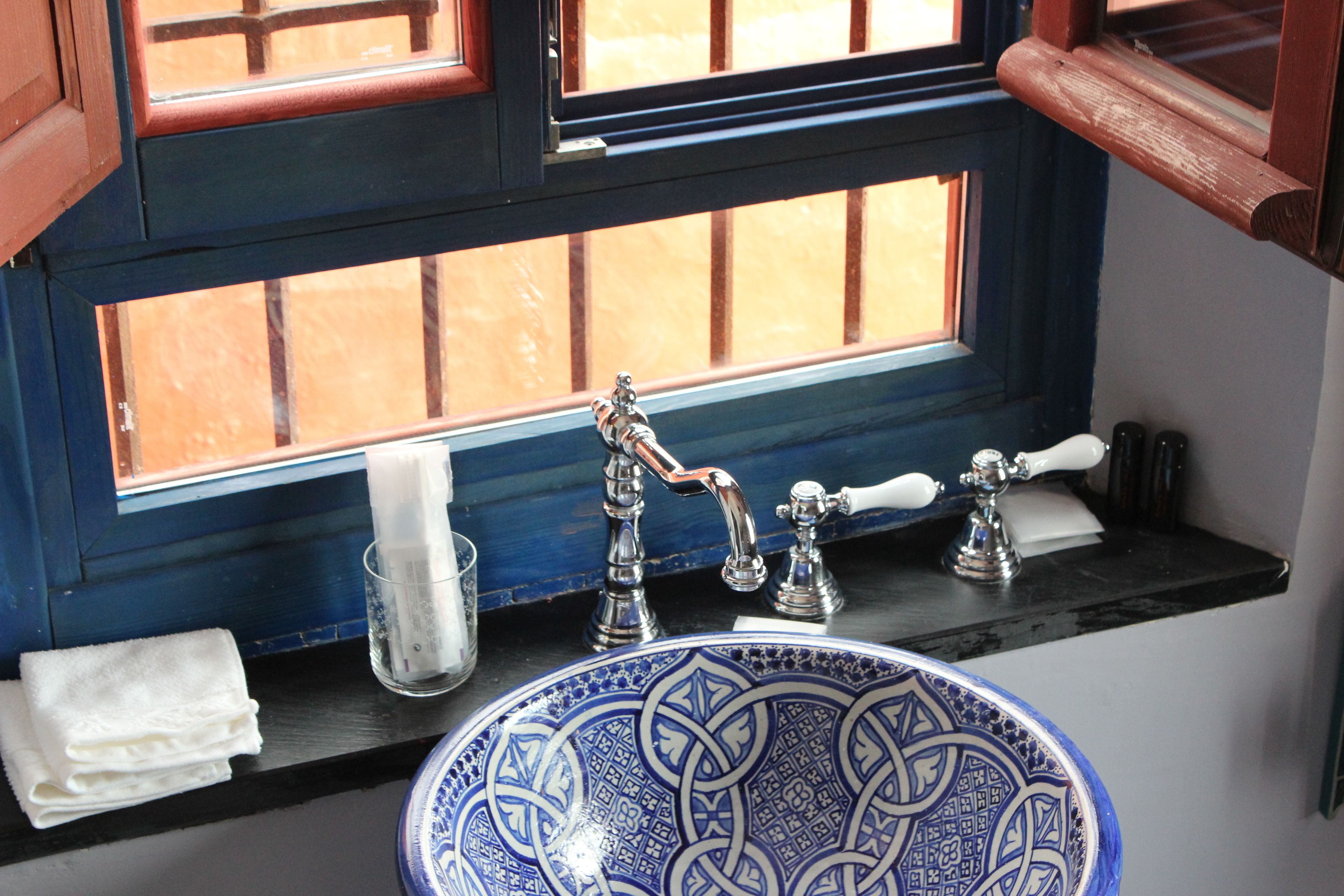 Een badkamer met een blauwe keramieken wasbak, een raam met houten kozijnen en een handdoek op het aanrecht.