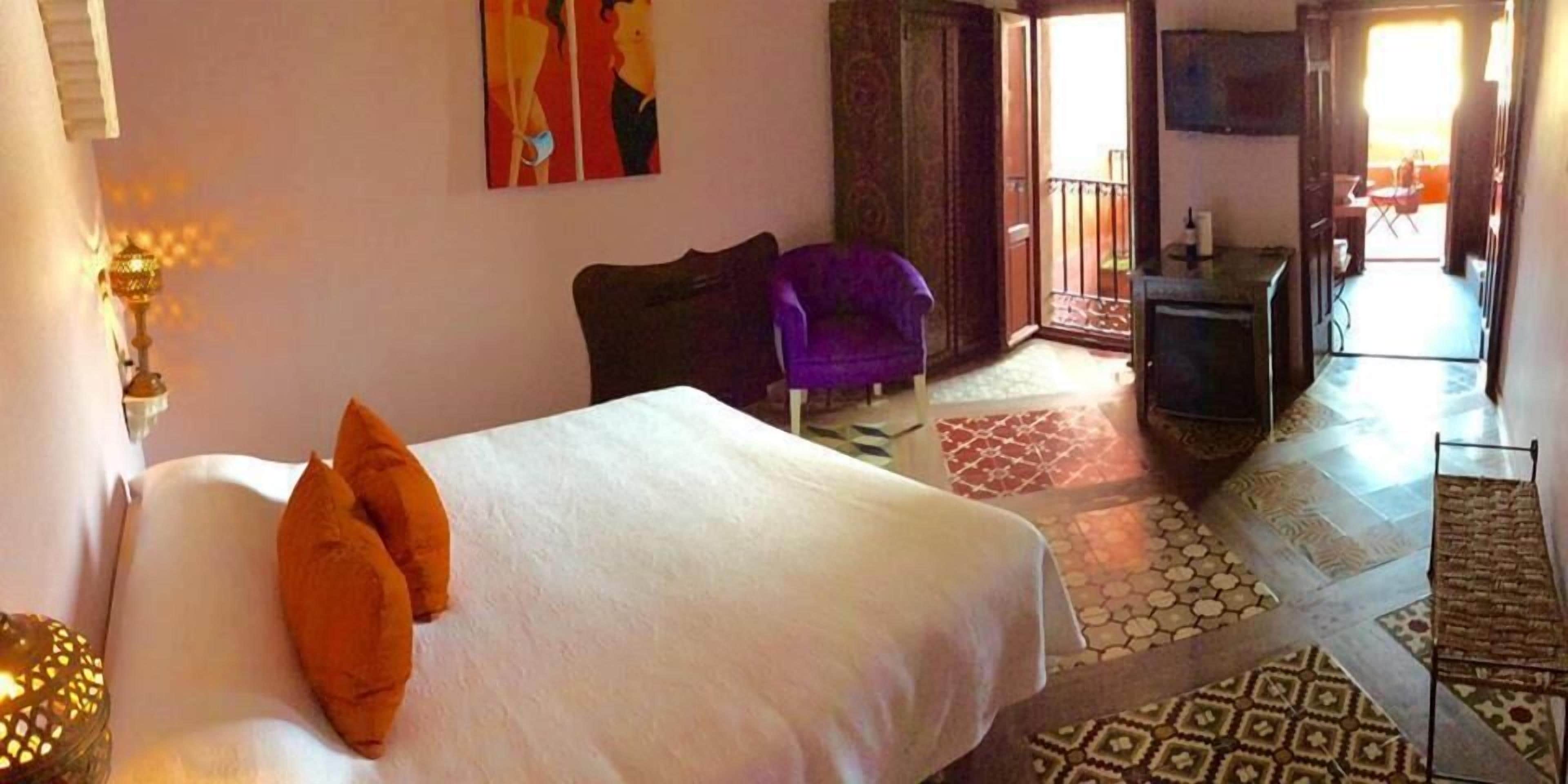 Una habitación de hotel con cama, una silla morada, un escritorio y un balcón.