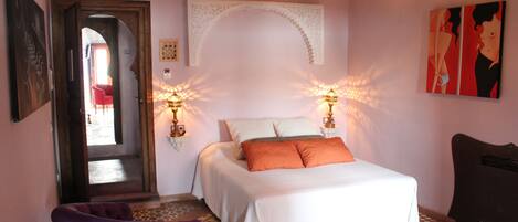 A bedroom with a bed, two bedside lamps, a chair, a door, and a wall with artwork.