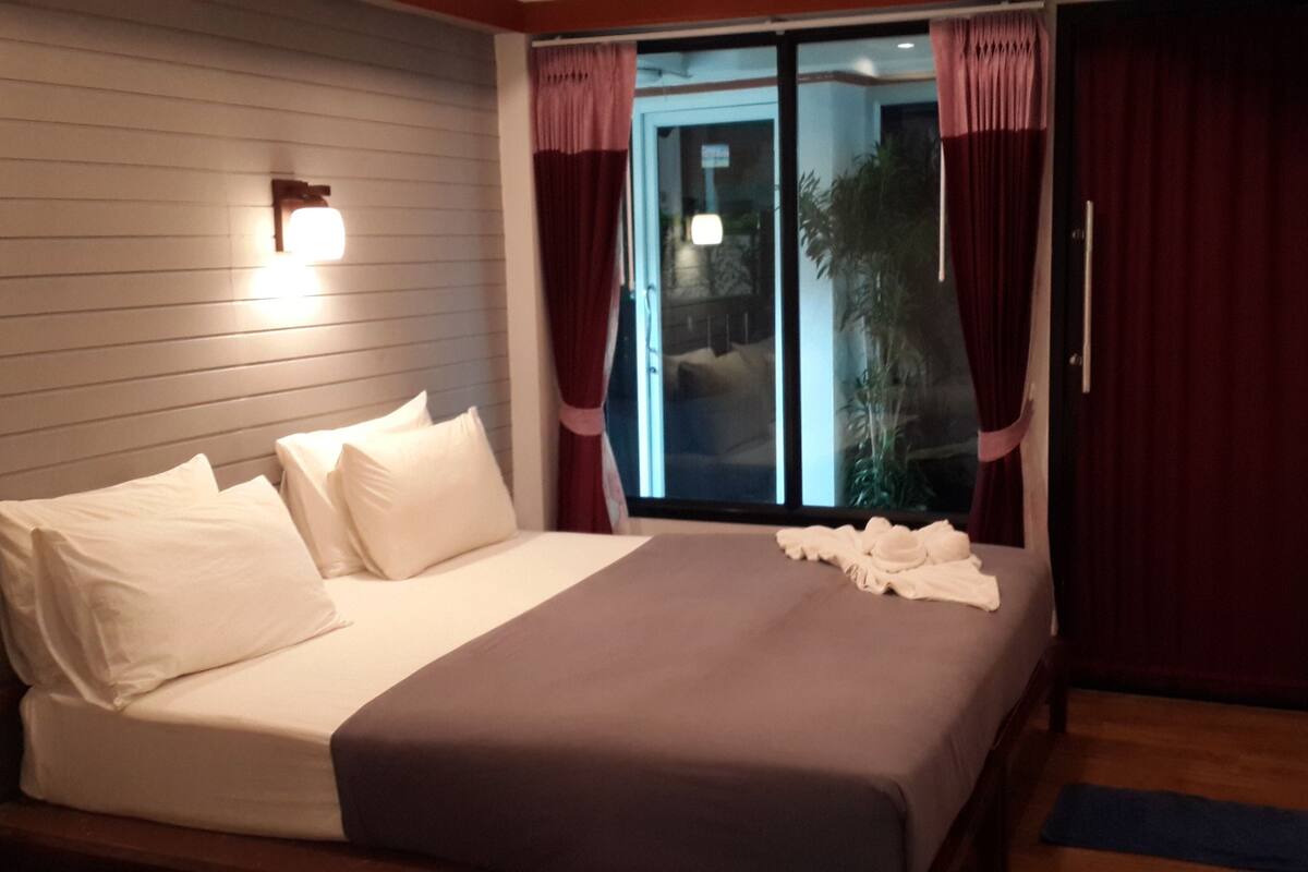 A bedroom with a bed, pillows, a nightstand, a window with curtains, and a view of the outdoors.