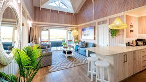 A spacious living area with a large mirror, wooden flooring, a sofa, a coffee table, and a kitchen area with a bar and stools.